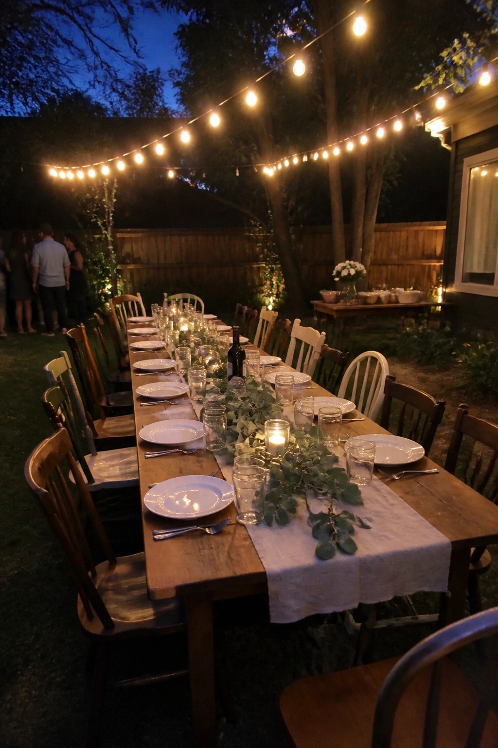 Long wooden table set with white plates and greenery under glowing string lights in a backyard at twilight for a wedding dinner