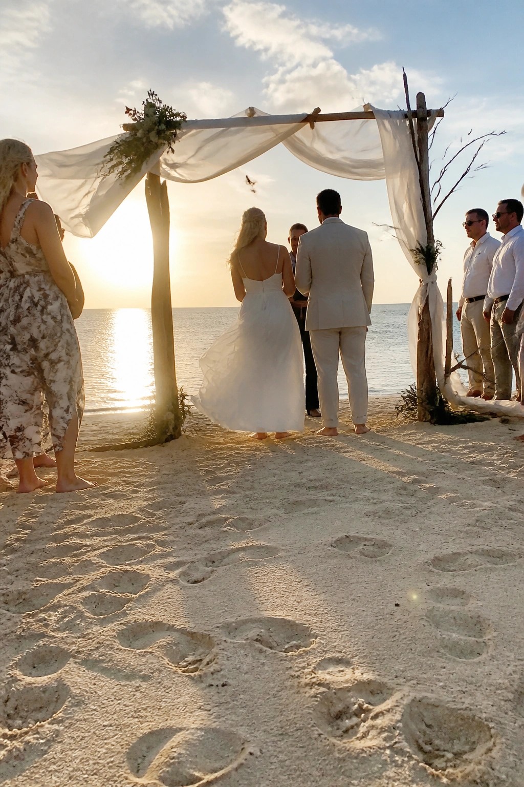 A bride and groom stand barefoot under a white draped wooden arch on a sandy beach at sunset facing the ocean with footprints visible in the foreground.