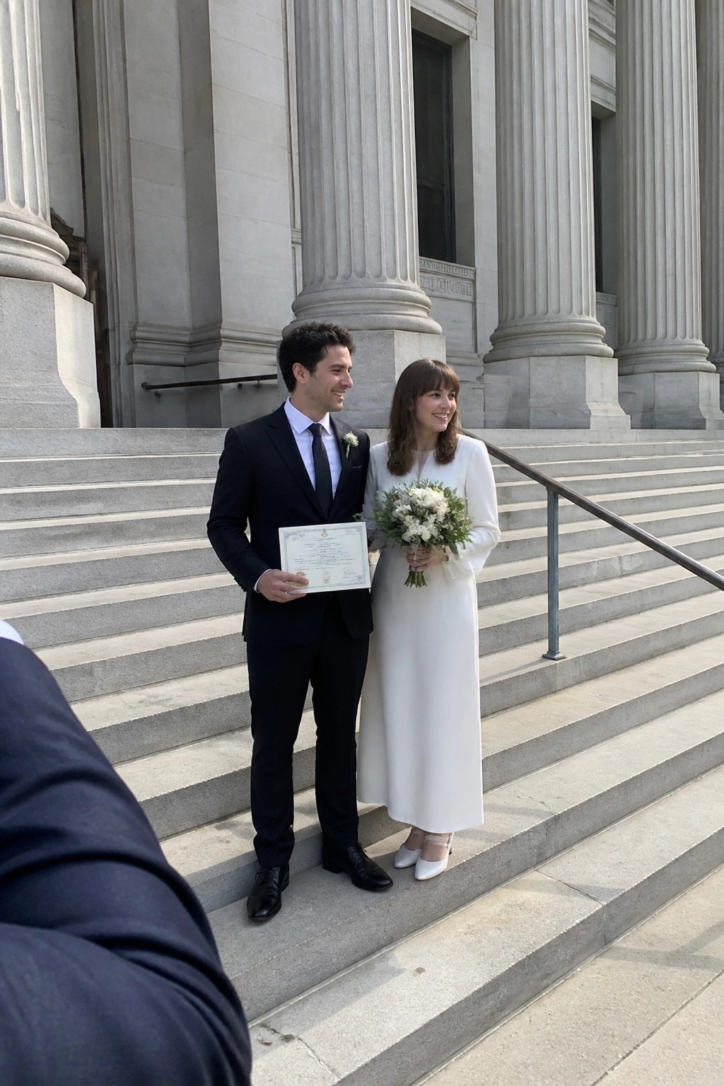 Bride in white dress and groom in black suit smile on stone steps of columned building while holding marriage certificate and white flower bouquet