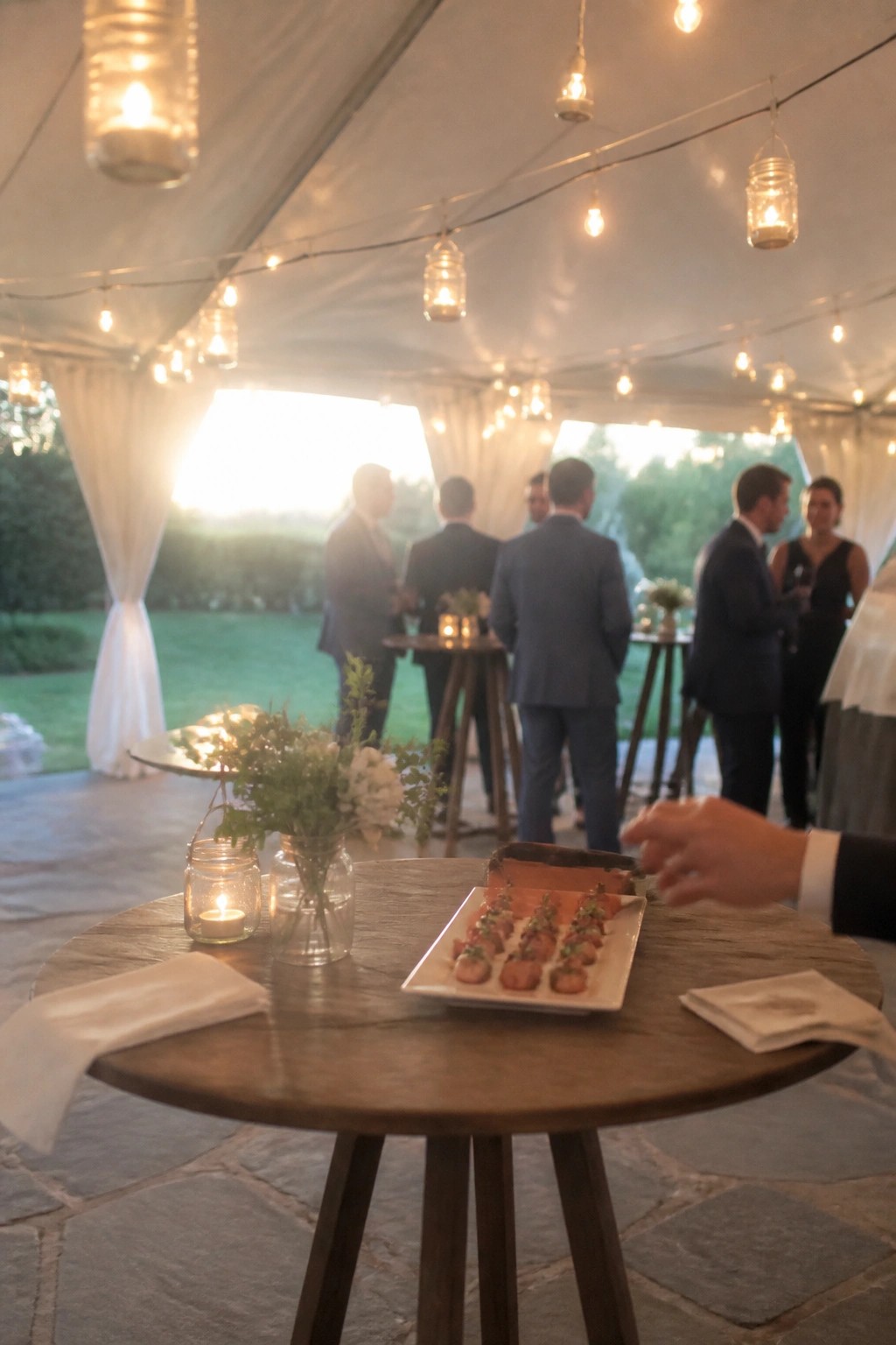 Wooden table with appetizers and candlelit jars inside a tented wedding space featuring string lights and formally dressed guests at dusk