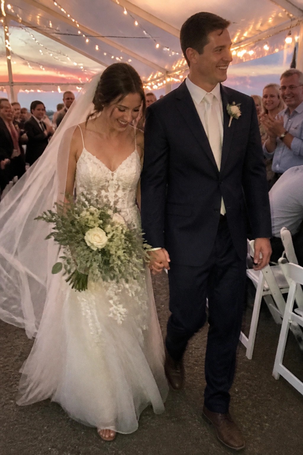 Bride in lace gown and groom in navy suit exit hand in hand under a tent with string lights at sunset while guests clap in the background.