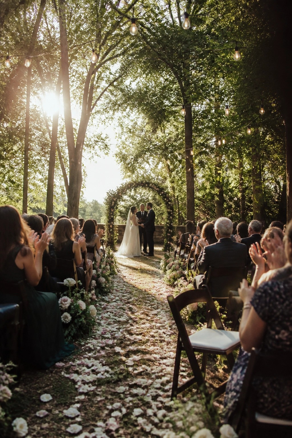 A wedding ceremony in a forest with guests on wooden chairs along a rose petal aisle string lights overhead and a couple standing under a floral arch as sunlight filters through the trees.