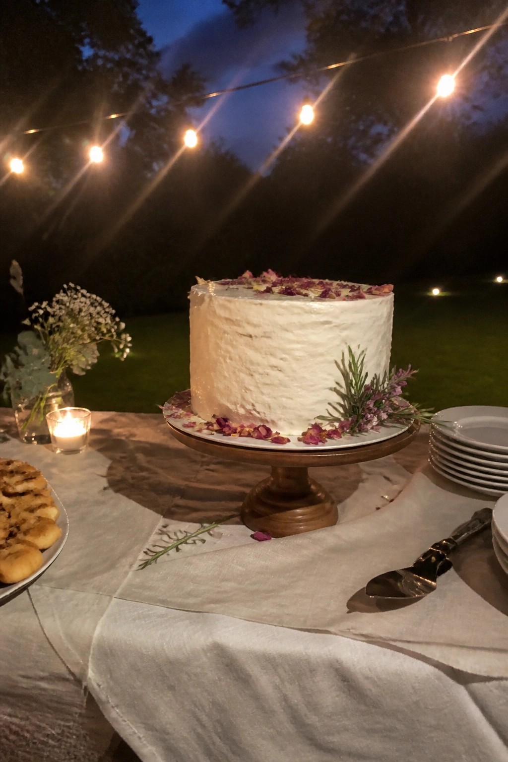 A white wedding cake with rose petals and herbs on a wooden stand outdoors at dusk under string lights with cookies and tableware on a linen cloth.