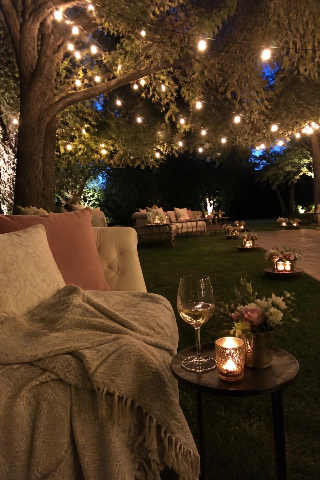 Cozy armchair with blanket, wine glass, and candle under glowing string lights in garden.