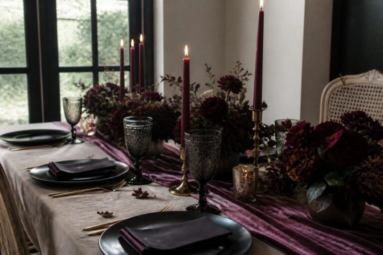 A moody wedding table with plum flowers, red candles in gold holders, black plates, and gold cutlery on a wine velvet runner over dark wood.