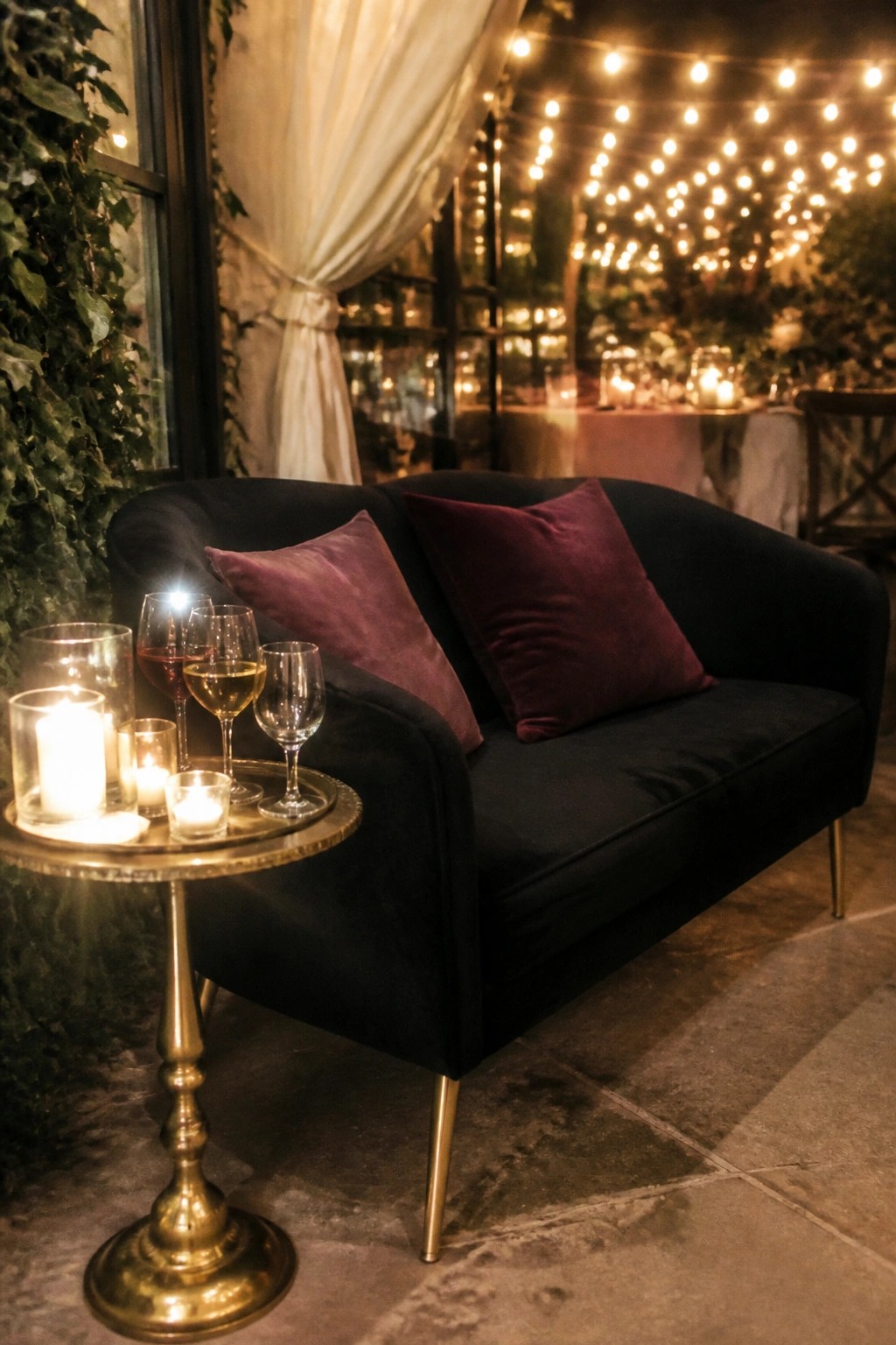 Black velvet sofa with plum pillows beside a gold table holding lit candles and wine glasses under string lights at a moody romantic wedding lounge.
