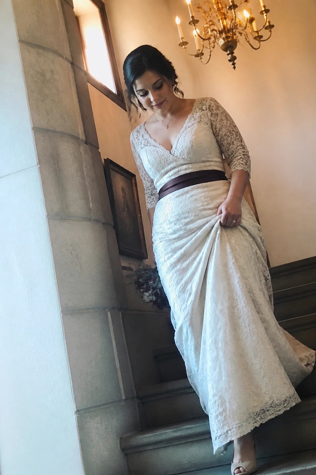 Bride in ivory lace wedding dress with wine sash stands on stone staircase under chandelier in moody romantic style