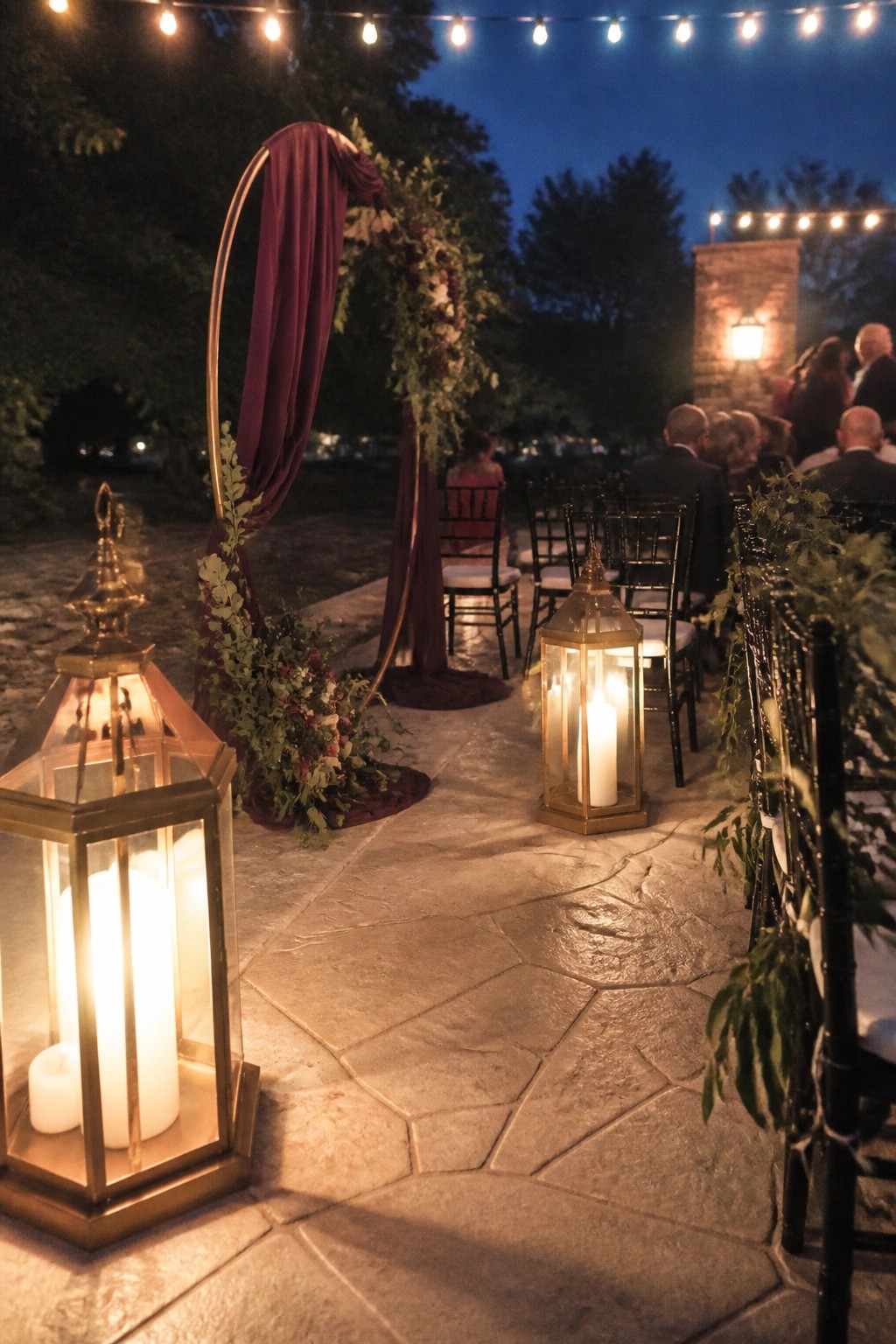 Nighttime outdoor wedding aisle with gold lanterns, plum draped circular arch, black chairs, and string lights on a stone path.