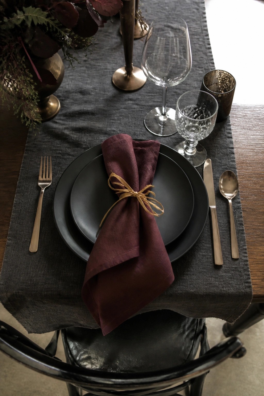 Dark romantic wedding table setting with black plates, wine colored napkin tied in antique gold ribbon, charcoal runner, gold cutlery, and moody florals in bronze vase.