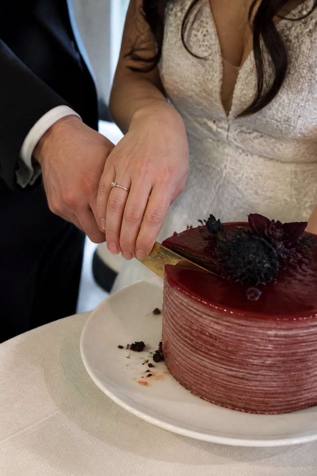 Bride and groom cutting a deep wine red layered cake with gold knife and dark floral accents in a moody romantic style.