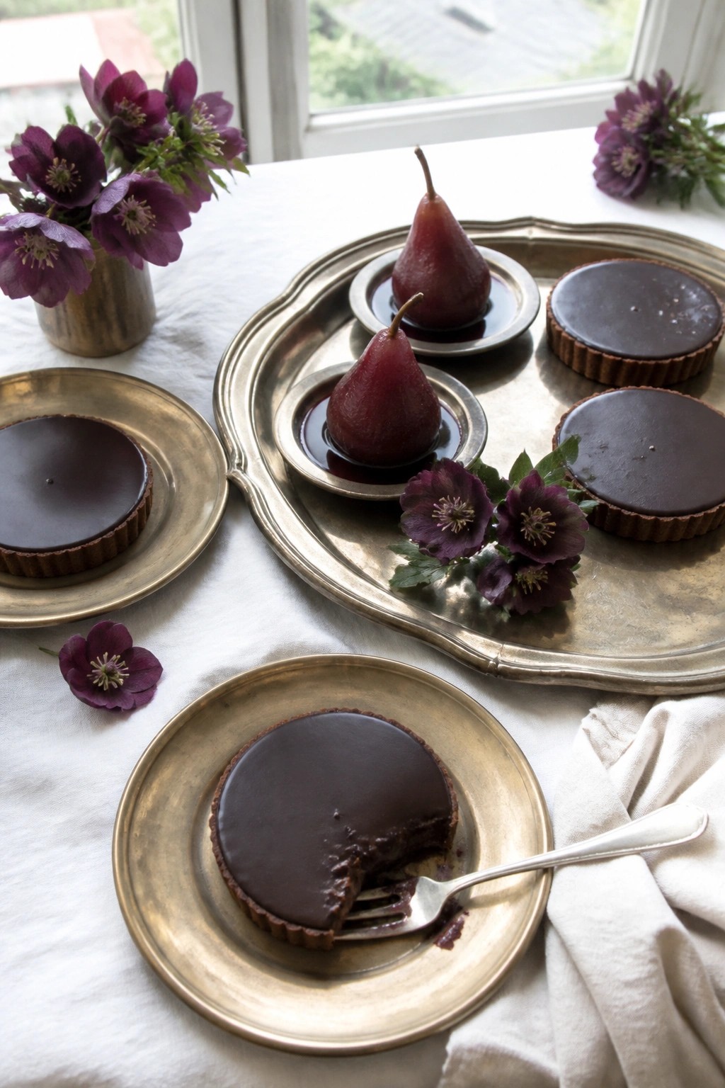 Moody wedding dessert display with dark chocolate tarts, wine poached pears, and plum flowers arranged on antique gold trays and plates.