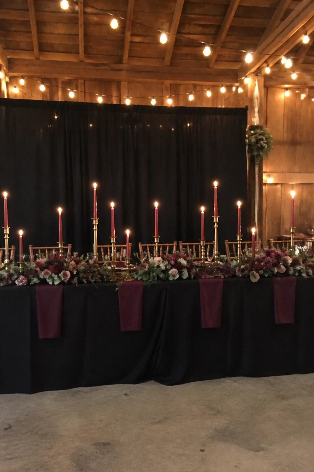 Moody wedding head table with black tablecloth, gold candlesticks holding lit red candles, plum floral garlands, and wine napkins in a rustic barn with string lights and black backdrop.