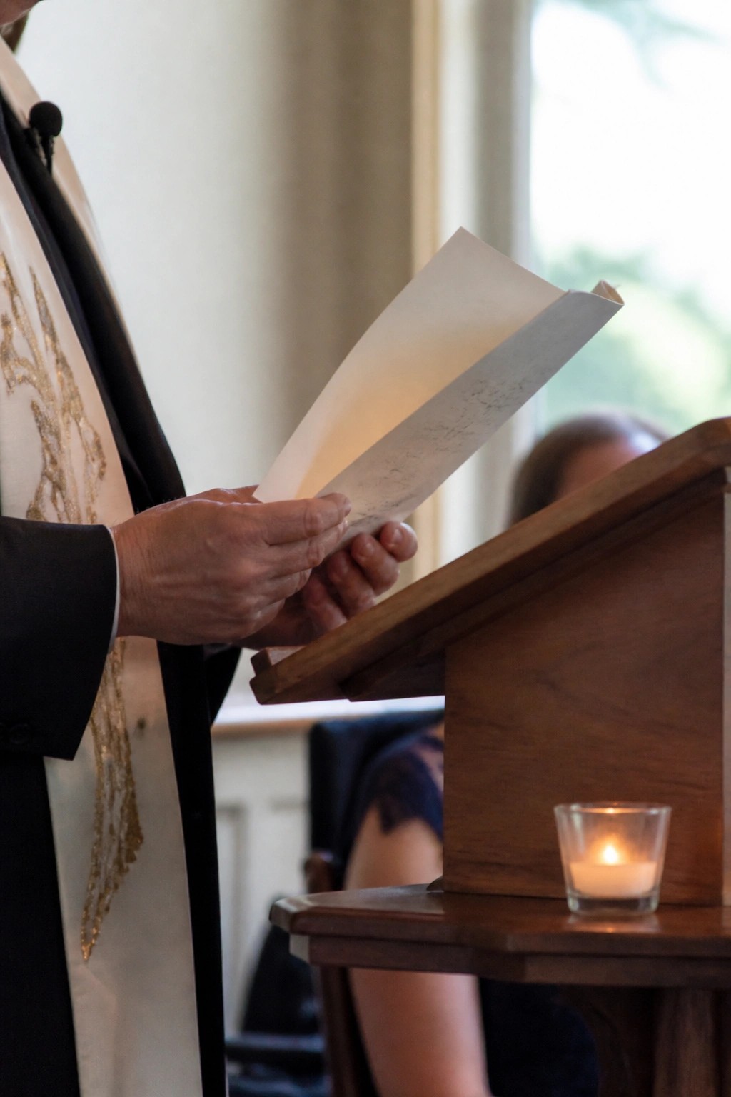 Clergyman in ornate white and gold vestments reads handwritten papers at a wooden podium with a lit candle nearby during a wedding ceremony.