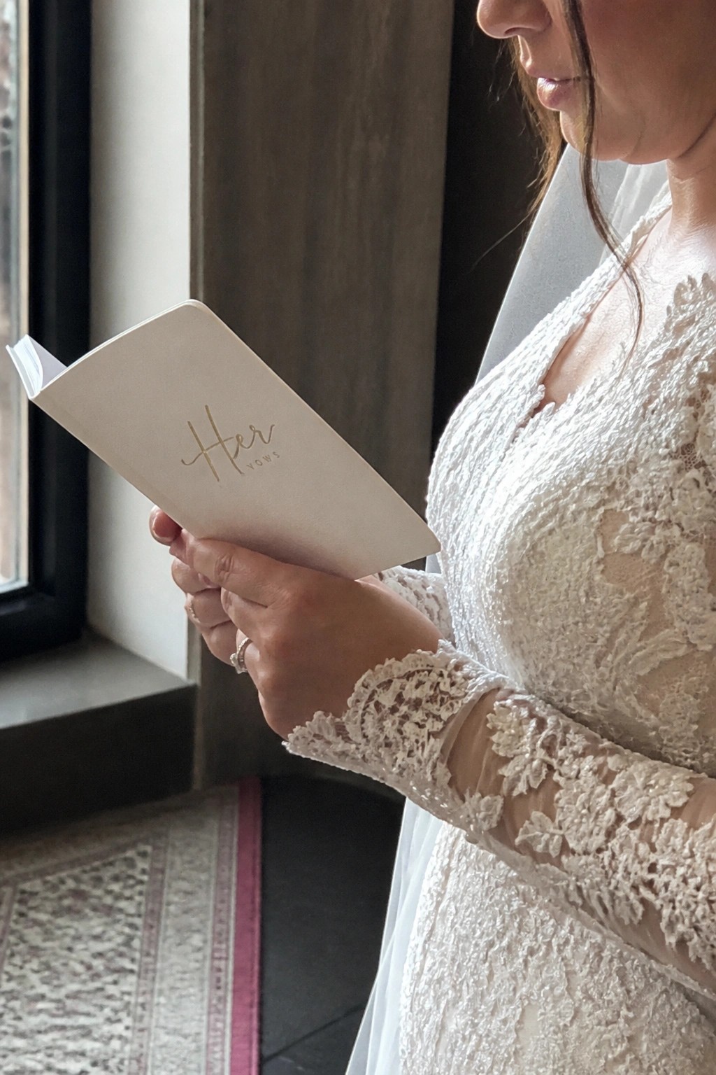 Bride in white lace dress reads from a small vow book labeled Her Vows in gold lettering while standing by a window.