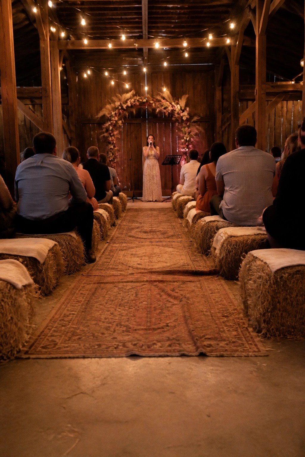 Bride in lace gown stands under floral arch at end of rug aisle in rustic barn with guests seated on hay bales and string lights overhead