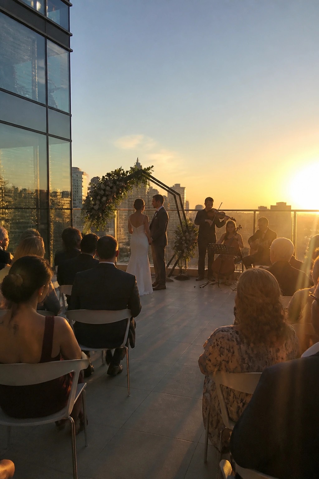 Bride and groom stand under a floral arch on a rooftop at sunset while a string trio performs with a city skyline in the background