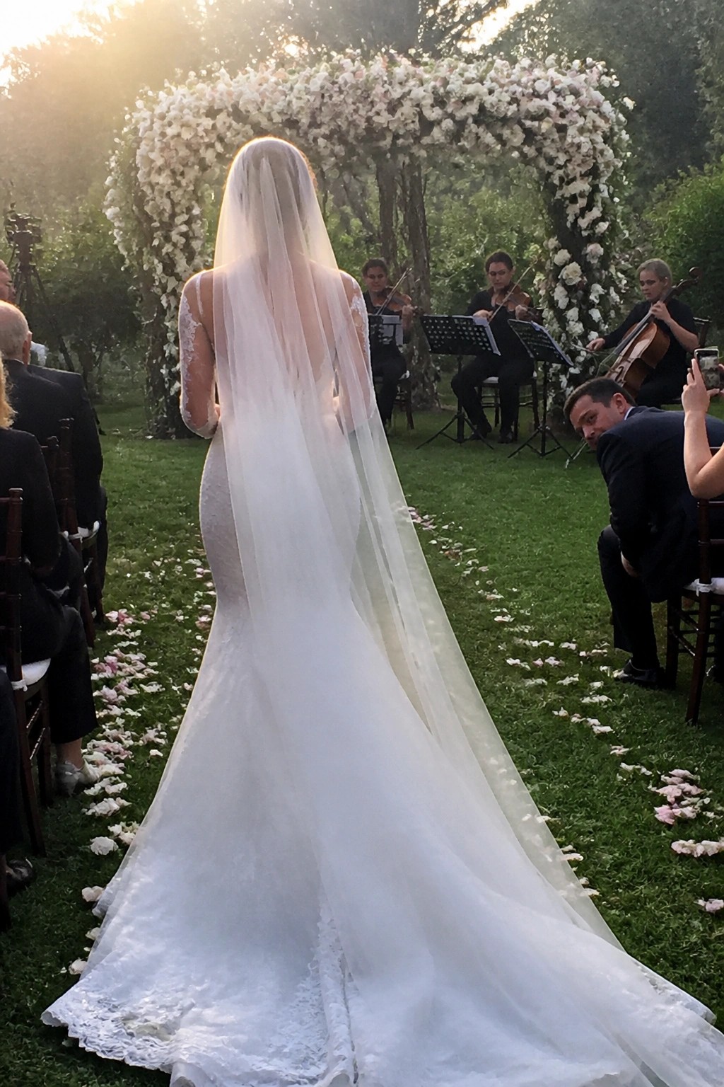 Bride in lace gown and long veil walks toward a large white floral arch while a string quartet performs at an outdoor garden wedding during golden hour.