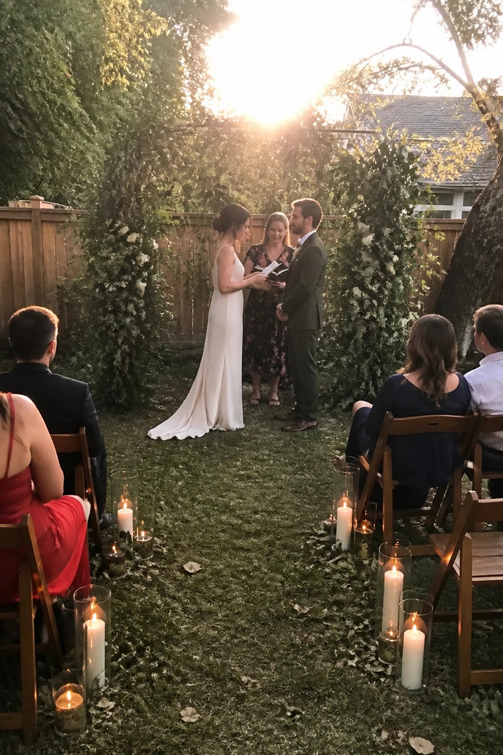 Outdoor wedding ceremony at sunset with bride in white gown and groom in green suit under floral arch candles lining the grass aisle among seated guests