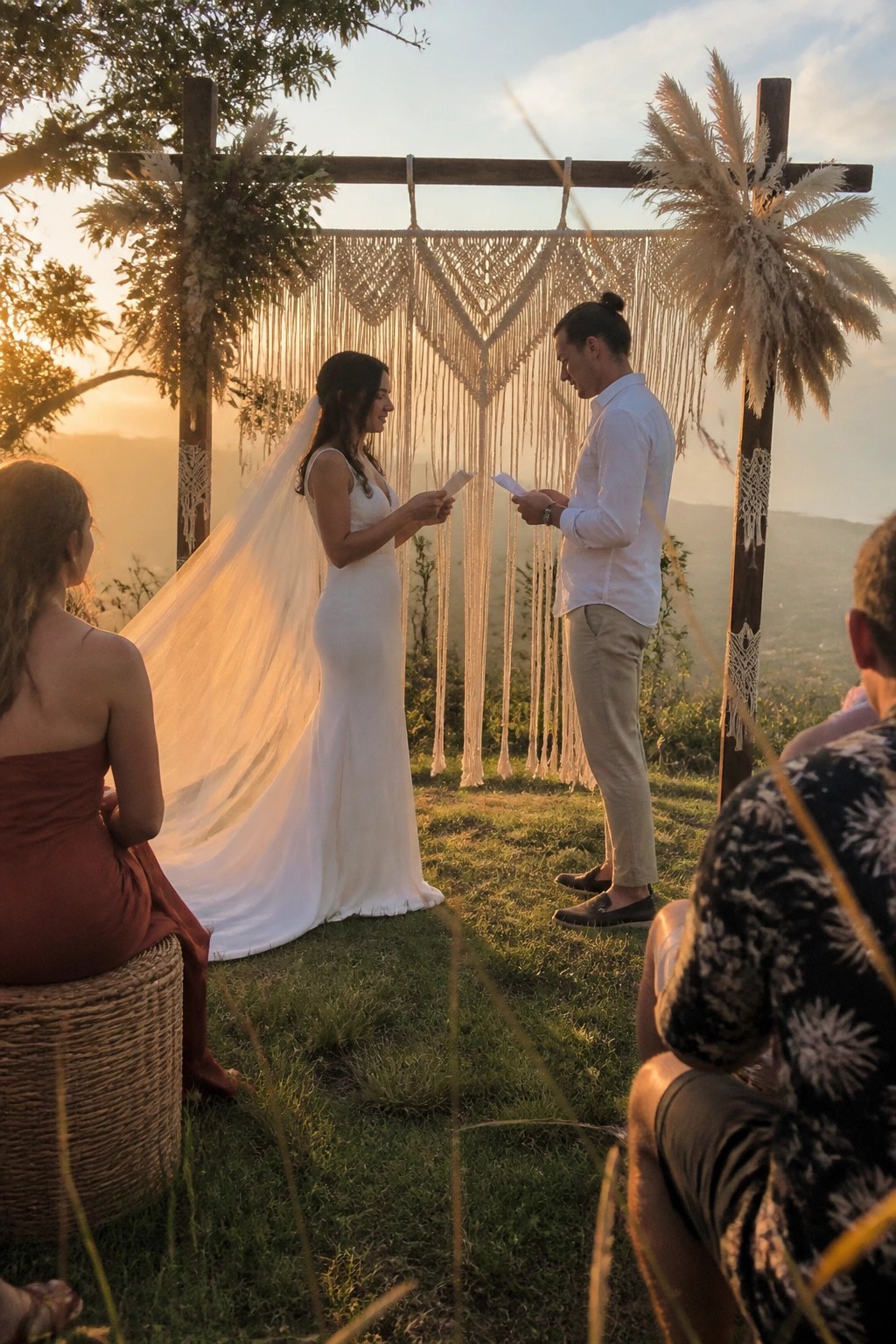 Bride and groom read vows under a macrame and pampas grass arch at sunset with guests seated on grass in a mountain landscape.