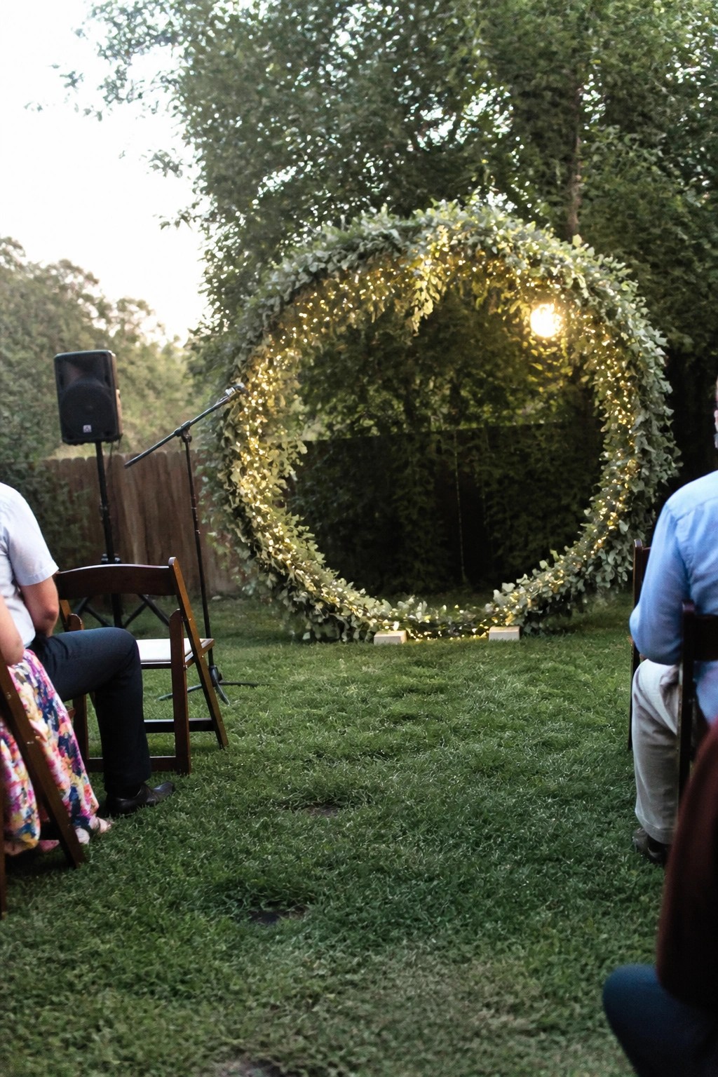 Outdoor wedding ceremony with circular arch of greenery and fairy lights on grass at dusk guests seated on chairs microphone and speaker nearby trees in background
