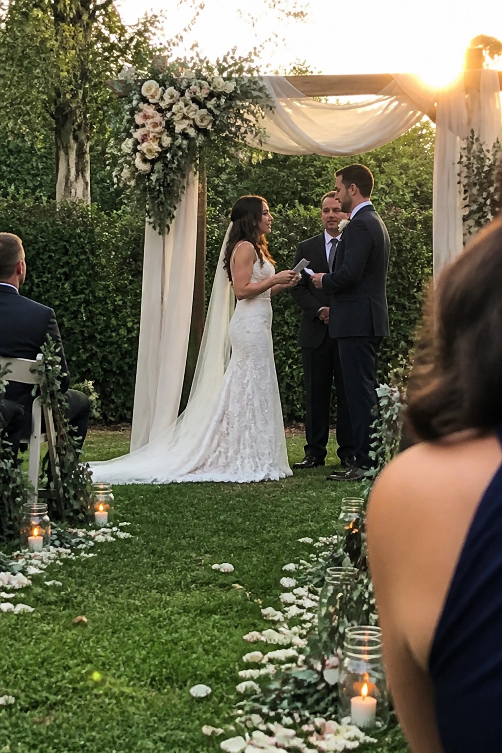 Bride and groom exchange vows under a wooden arch covered in white roses and fabric on a petal strewn lawn at sunset