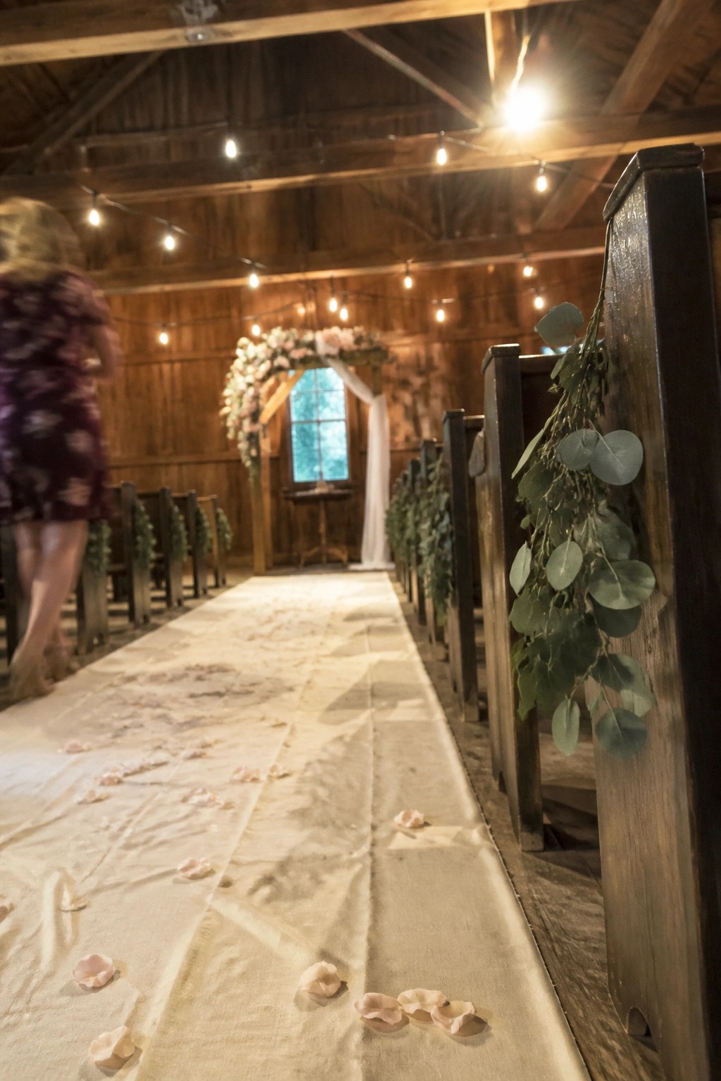 Rustic barn wedding aisle with wooden pews draped in eucalyptus, pink petals on a cream runner, string lights above, and a floral arch at the end.