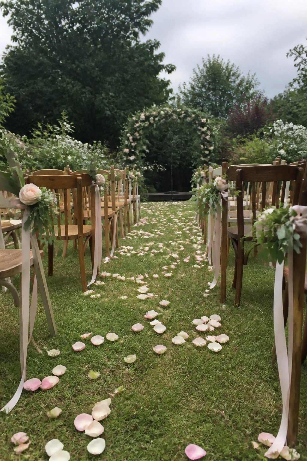 Outdoor wedding ceremony on grass with wooden chairs decorated in pink flowers and ribbons and a petal covered aisle leading to a floral arch.