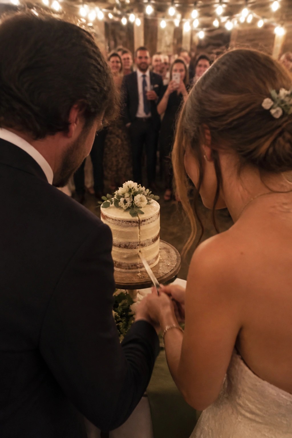 Bride and groom cutting a white floral wedding cake from behind in a rustic venue with string lights and guests watching.