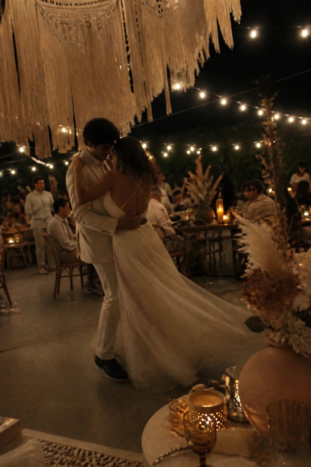 Bride and groom slow dancing at night under macrame hangings and string lights with pampas grass decor at an outdoor boho wedding reception