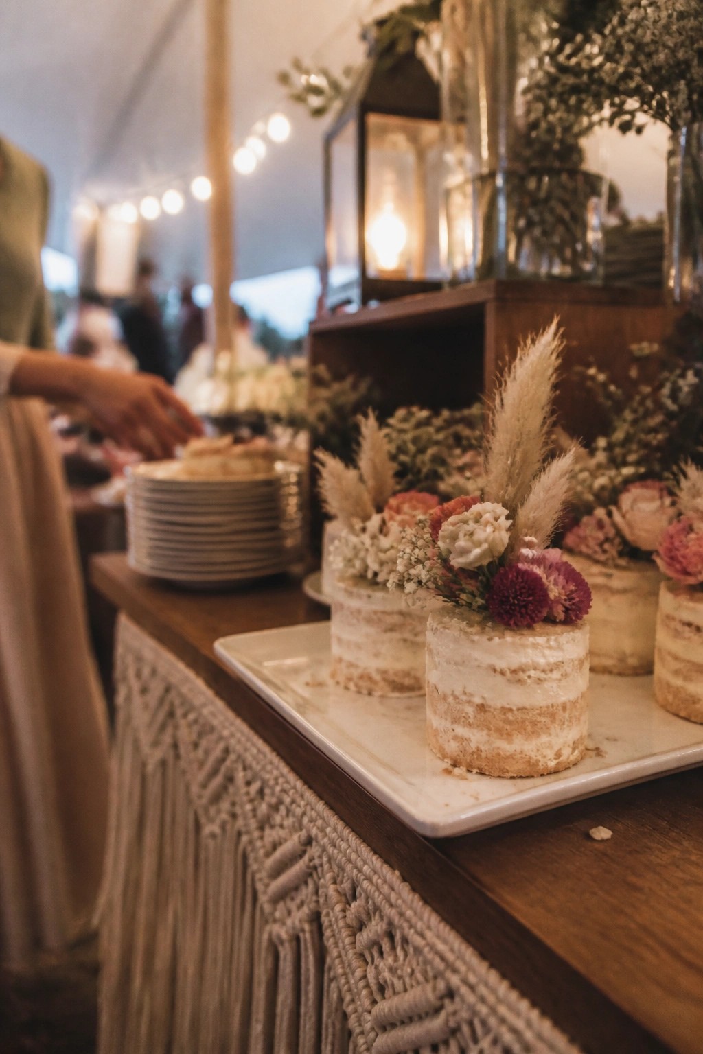 Small naked wedding cakes topped with pampas grass and mixed florals sit on a white tray atop a wooden table with a macrame runner in a warmly lit boho reception space.