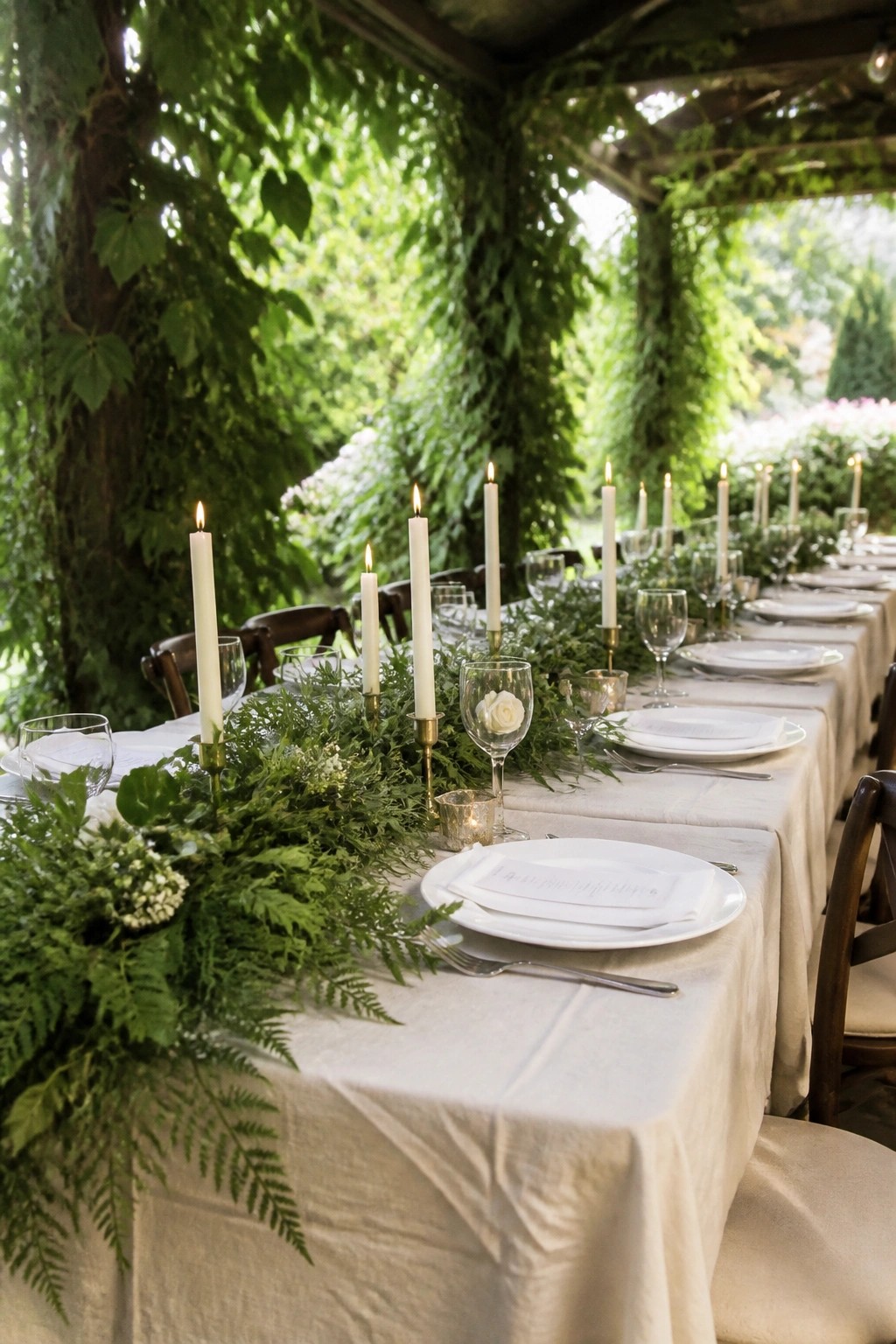 Long outdoor wedding table with white linens, lit candles, and lush green foliage runner under a vine covered pergola in a garden setting.