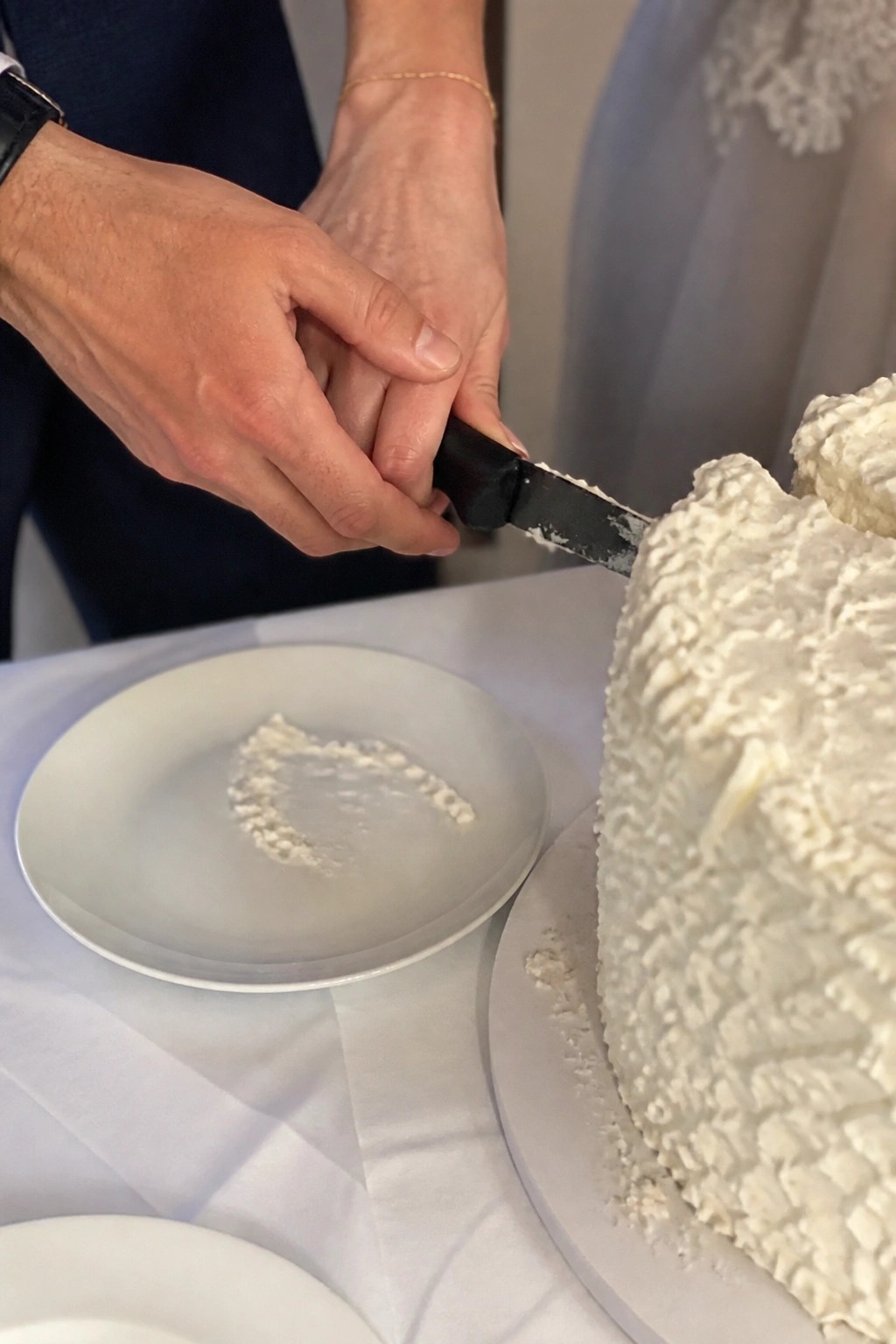 Close up of hands cutting a white wedding cake with a black knife at a formal reception with a white plate nearby.
