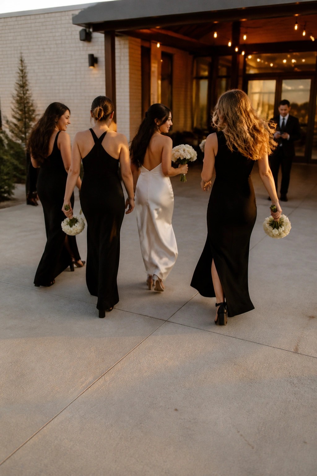 Bride in white satin gown leads bridesmaids in black dresses holding white bouquets as they walk toward a modern venue at dusk