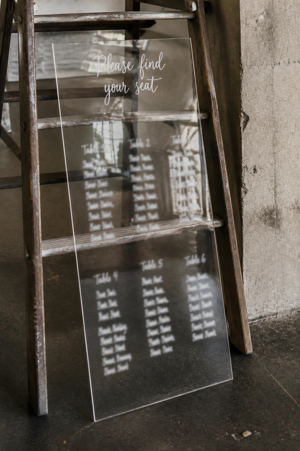Transparent acrylic wedding seating chart with table assignments displayed on a rustic wooden ladder against a concrete wall.