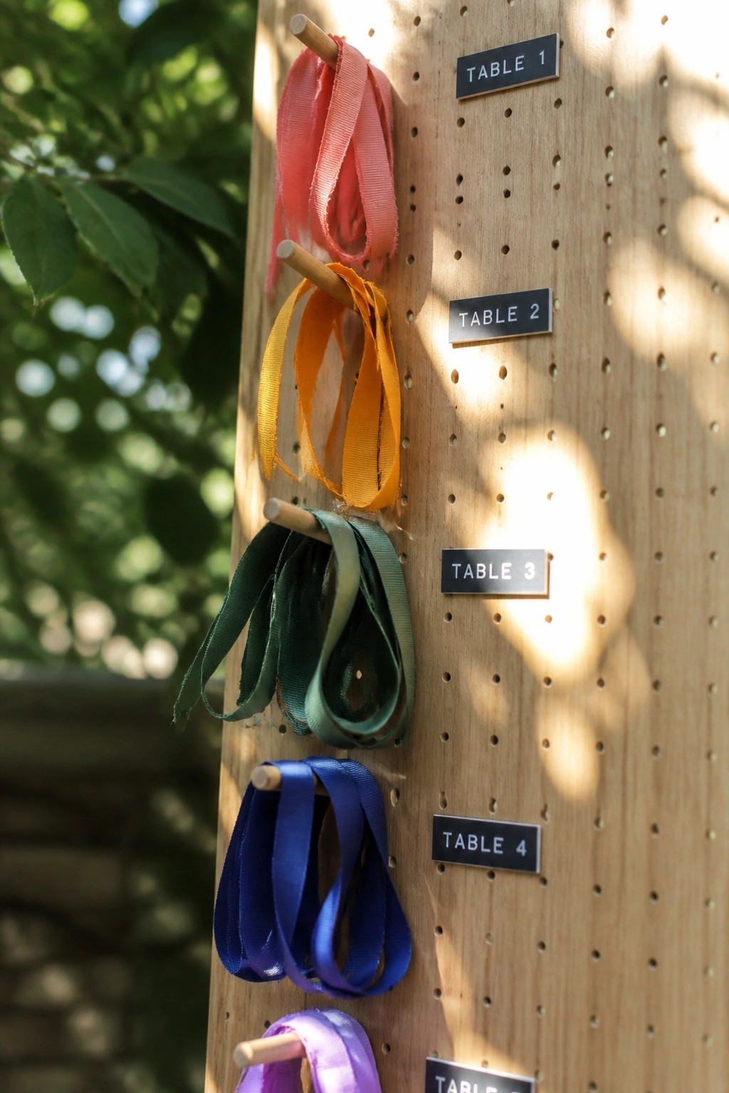 Wooden pegboard with pink orange green blue and purple ribbons hanging from pegs next to table number signs in an outdoor setting with sunlight and foliage