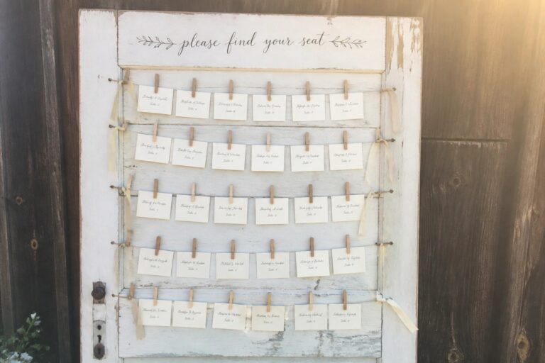 21 Clever Wedding Seating Ideas That Help Guests Find Their Tables and Feel Comfortable Rows of white place cards clipped with wooden pins on strings across a distressed blue vintage door, accented with pink ribbons for a rustic wedding seating display.