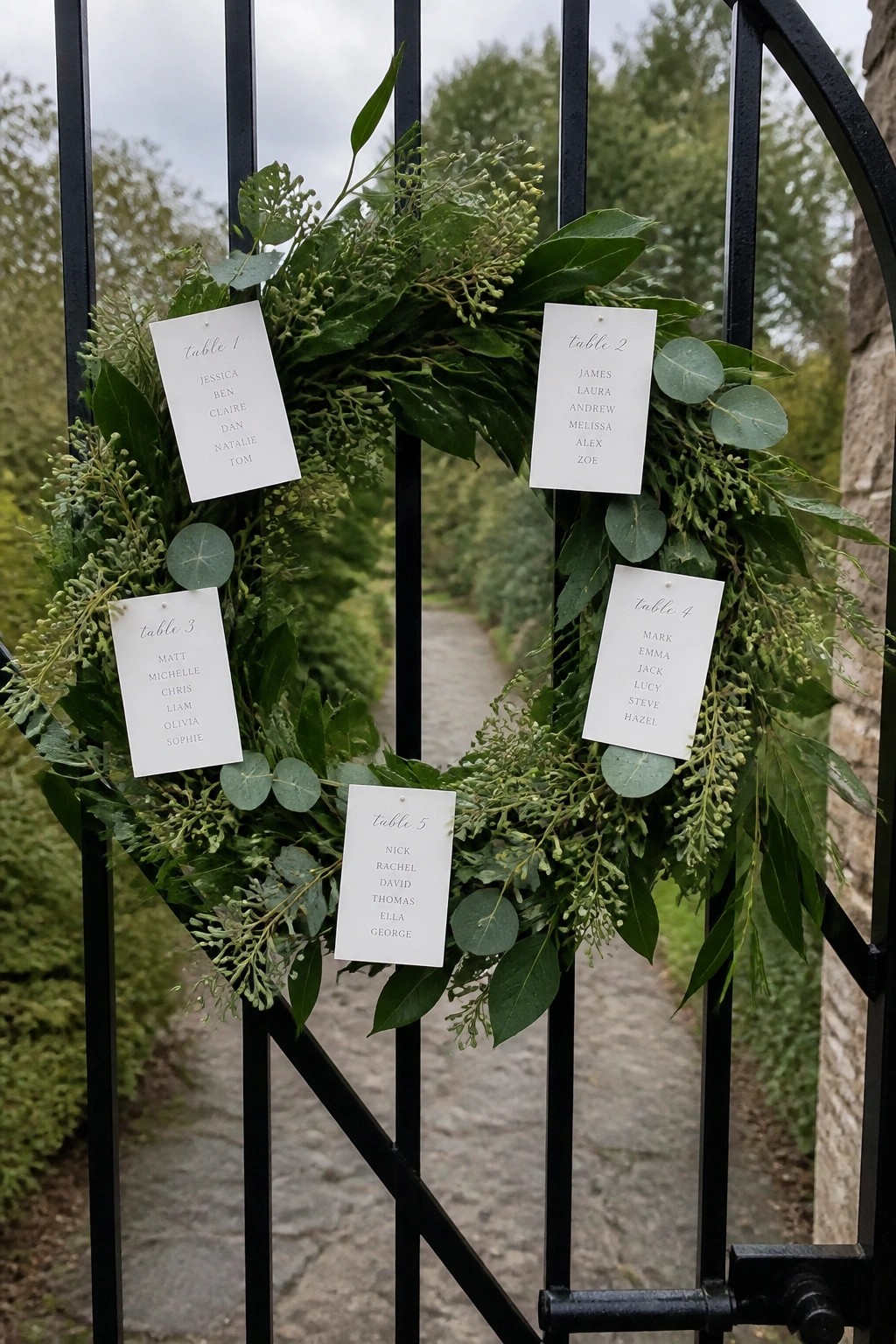 Black metal gate decorated with a lush green eucalyptus wreath displaying five white cards with wedding table assignments and guest names.