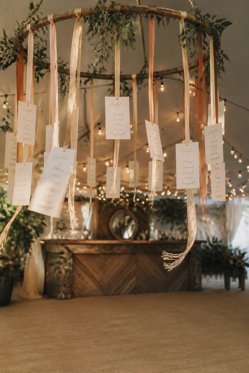A circular wooden hoop decorated with greenery and hanging peach and ivory ribbons holding white calligraphy seating cards in a warmly lit wedding reception venue.