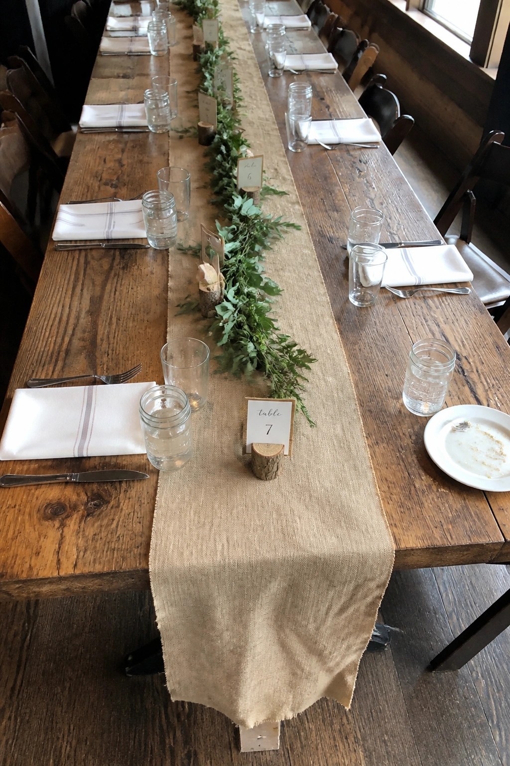 Long rustic wooden wedding table with burlap runner, green foliage garland, and wooden table number signs in a barn venue.