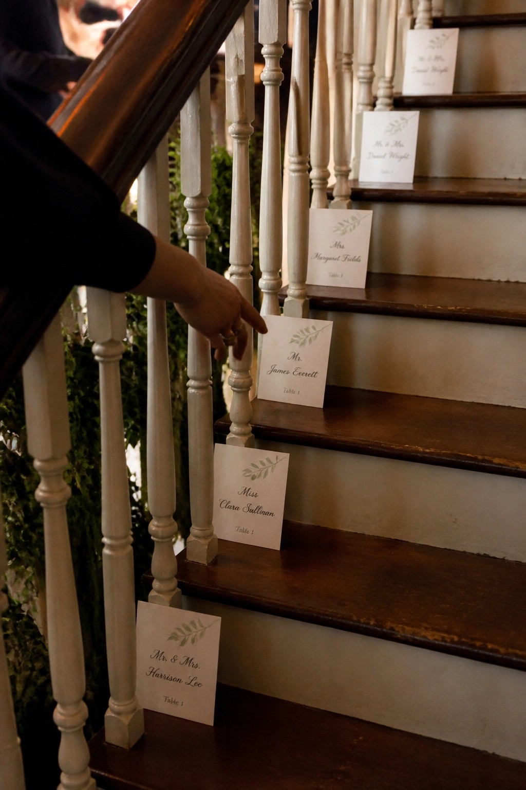 A wooden staircase with white balusters displays white place cards with leaf motifs listing guest names and table assignments for a wedding. A hand points to one card on the steps.