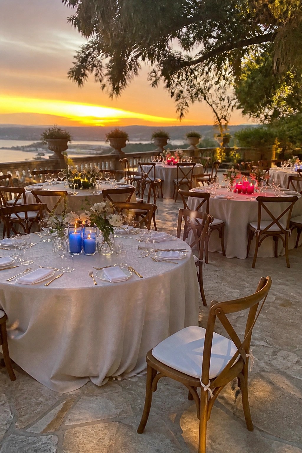 Outdoor wedding reception at sunset with round tables covered in white linens, wooden chairs, blue and pink candles, and floral centerpieces on a stone terrace overlooking hills and water.