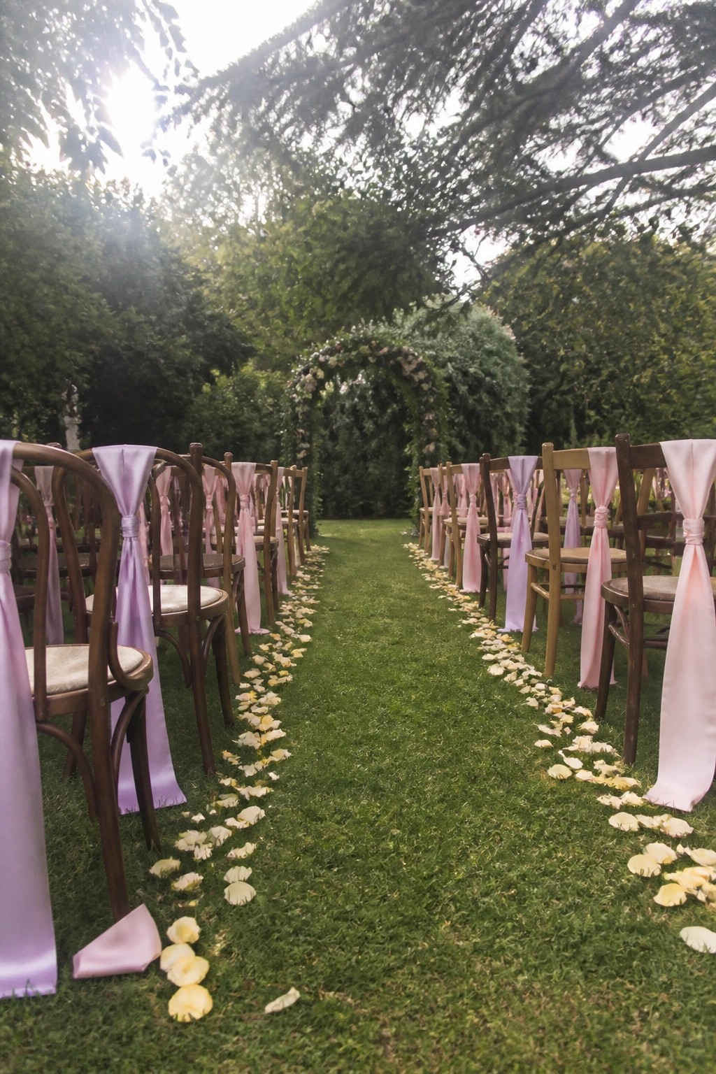 Outdoor wedding aisle with wooden chairs tied in lilac and blush ribbons yellow flower petals on green grass and a floral arch under leafy trees in soft sunlight