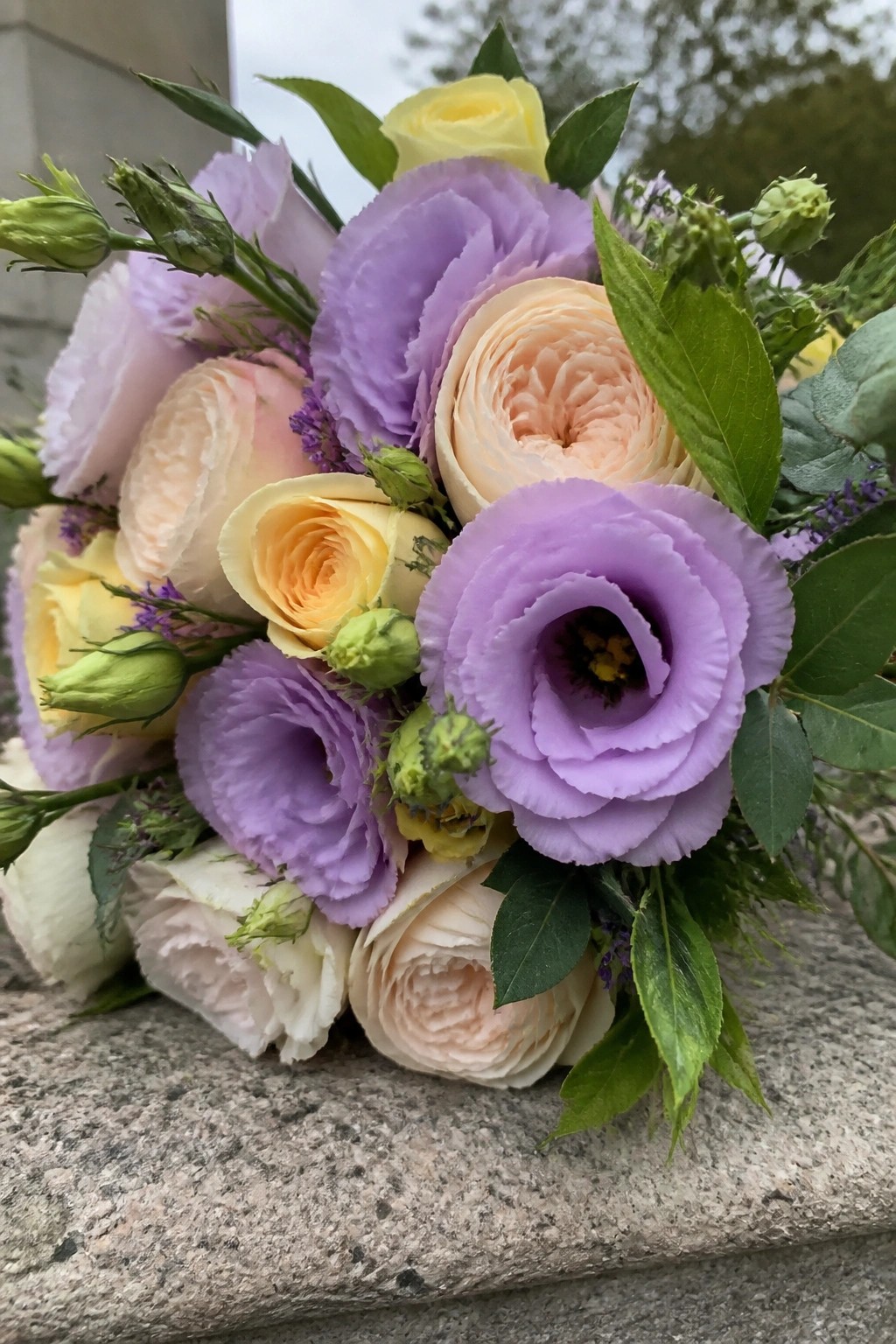 Close up of a spring wedding bouquet with lilac lisianthus, peach ranunculus, yellow roses and green foliage resting on stone.