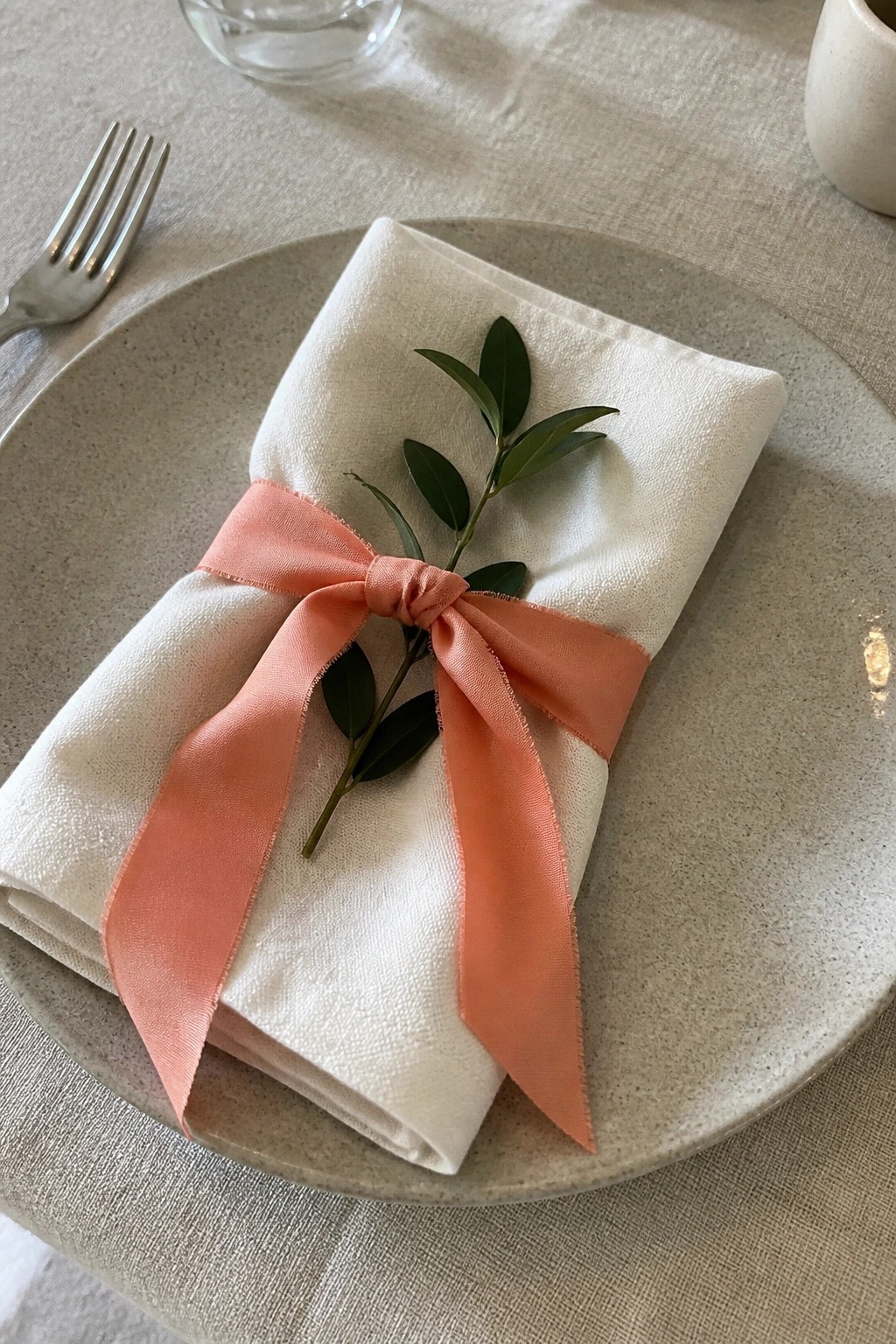 A white linen napkin folded on a beige plate is tied with a coral ribbon bow and accented with green leaves on a neutral table setting.