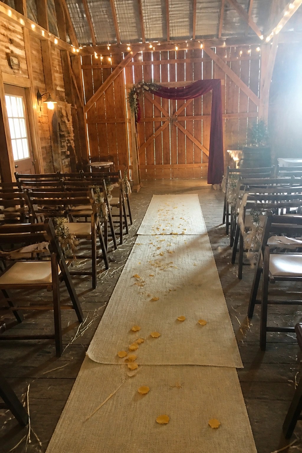 Rustic barn wedding aisle lined with wooden chairs and a burlap runner scattered with yellow petals leading to a maroon draped arch under string lights
