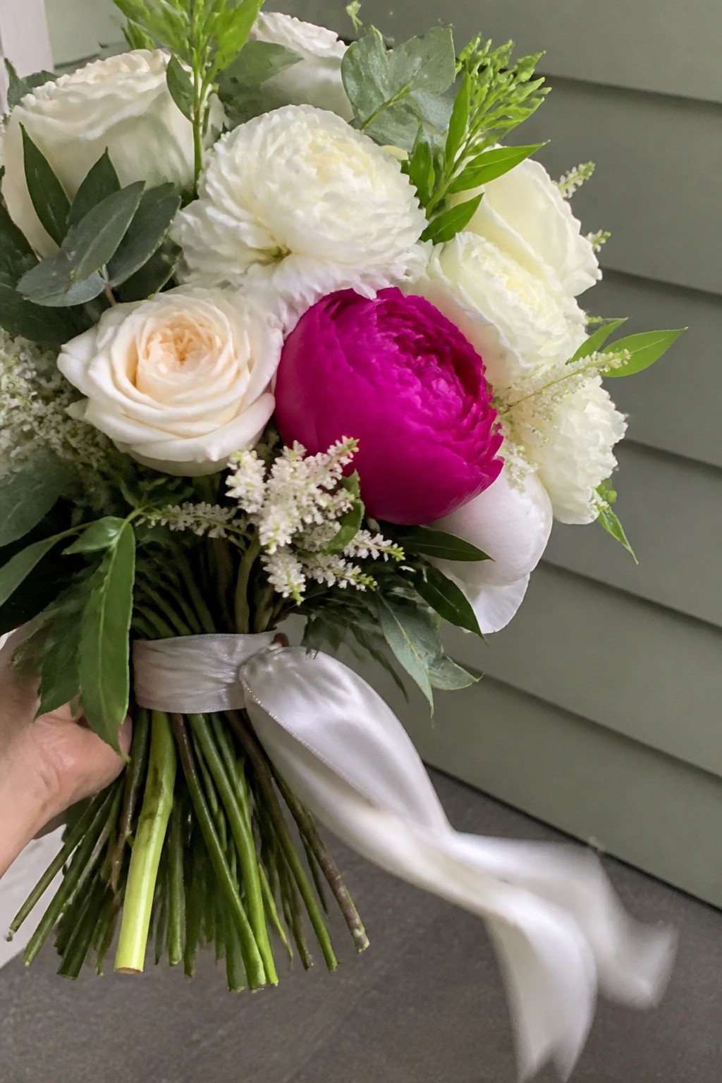A bridal bouquet of white roses and peonies with one bright magenta peony in the center tied by a white ribbon and held against a green wall