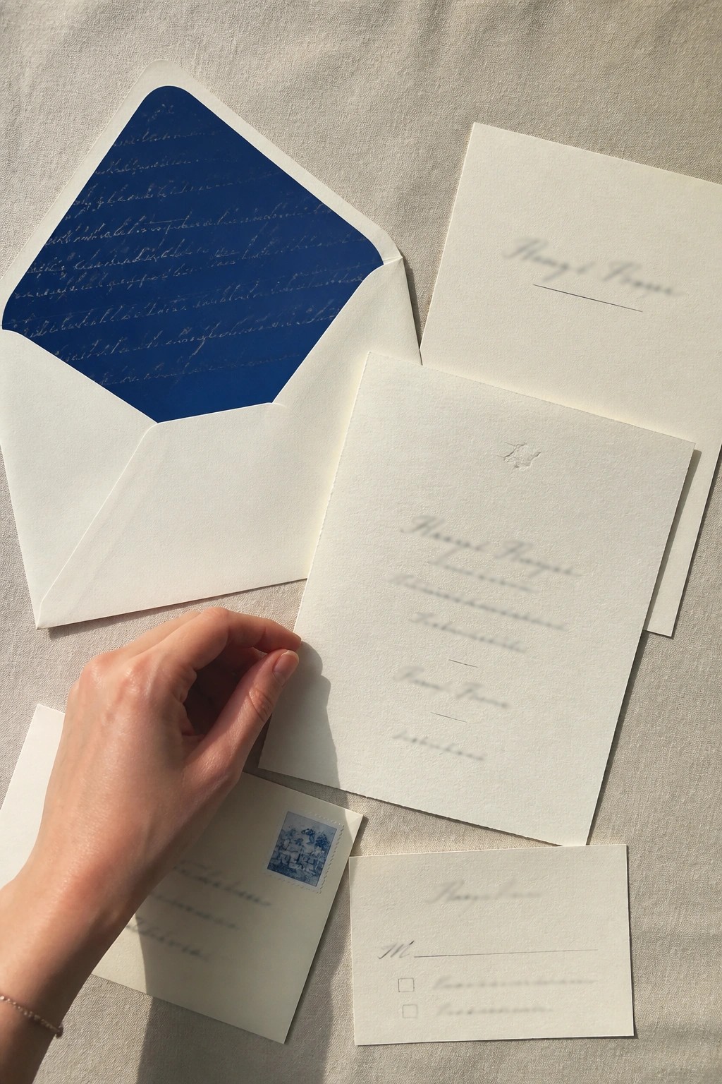 Elegant white and cream wedding invitations with bold blue envelope lining and stamp arranged on linen fabric with a hand holding one card