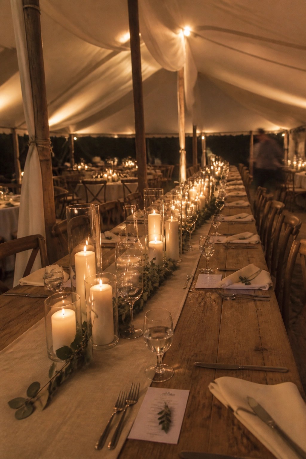 Long wooden wedding table set with glowing candles eucalyptus and rustic place settings inside a softly lit white tent at night