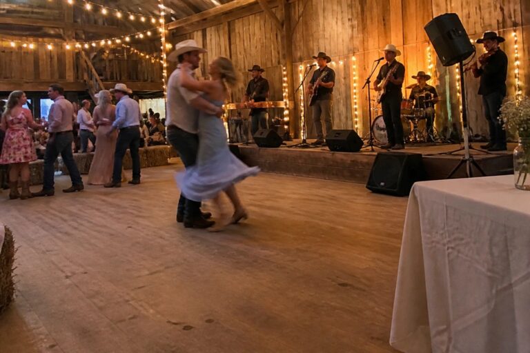 Bride in white dress dancing with partner in front of a live country band inside a wooden barn with string lights and guests on hay bales.
