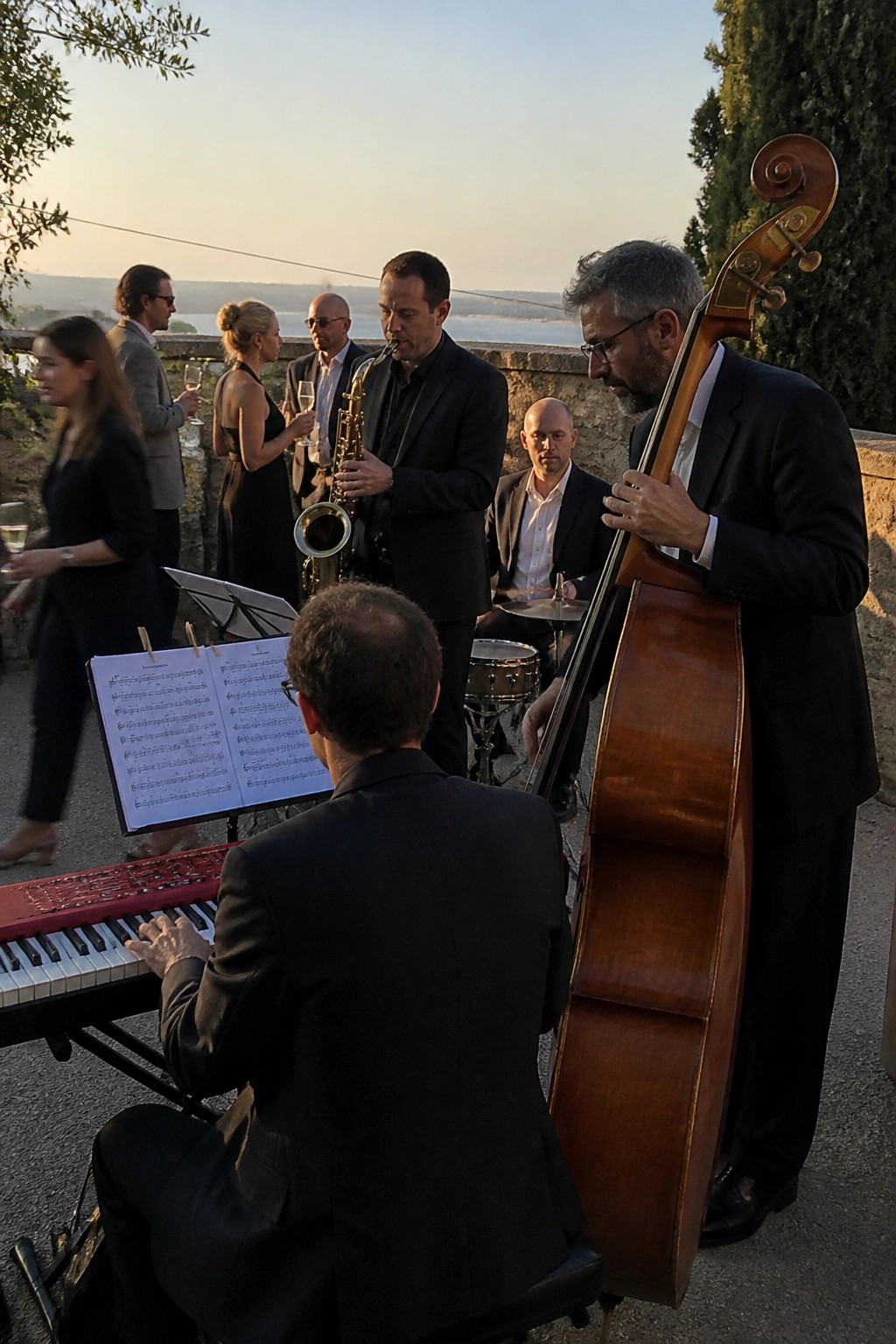 Jazz musicians in black suits perform at an outdoor wedding reception during sunset with guests mingling and a scenic landscape in the background.